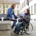 Das Bild zeigt zwei junge Frauen, die vor der Neuen Universität sitzen und sich unterhalten. Eine der Frauen sitzt in einem Rollstuhl und hält ein Buch in der Hand. 